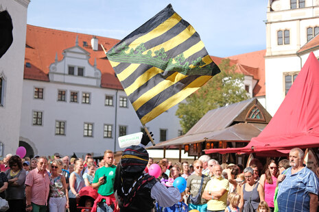 Eine sächsische Flagge weht über den Gästen eines Volksfestes.
